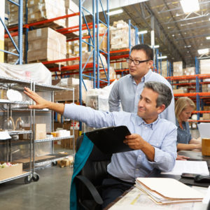 Business Colleagues Working At Desk In Warehouse Holding Clipboard