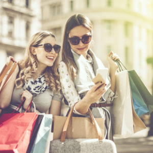 Cheerful beautiful women are shopping in the city. They are using smart phone, and one of them is holding a credit card.